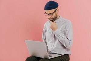 Photo of concentrated unshaven man holds chin, looks attentively at screen of laptop computer, enjoys online communication, wears optical glasses and formal outfit, isolated over pink background