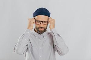 People, tension and migraine concept. Depressed unshaven young male suffers from headache, keeps hands in fists near temples, closes eyes, tries to concentrate, stands against white background photo