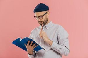 Photo of handsome male author has dark bristle, holds pencil and book, underlines necessary information, isolated over pink studio wall, has attentive gaze in notepad. Man notes something indoor