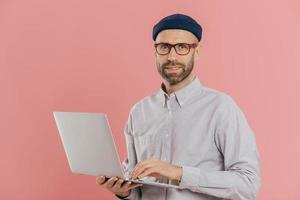Isolated shot of handsome unshaven male does distance job on portable laptop computer, wears hat and optical glasses, connected to wireless internet, stands over pink background, develops software photo