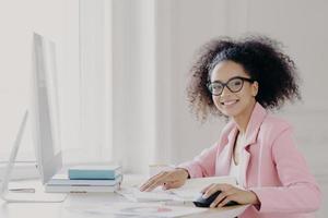 Prosperous curly female entrepreneur checks documents, studies contract, works on computer, wears glasses and pink jacket, poses at workplace, smiles broadly, happy almost finishing work in time photo