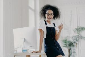 Happy carefree female financier uses mobile phone, glad to get salary in time, poses near wooden table, uses computer for work, wears white t shirt and sarafan, texts message, smiles pleasantly photo