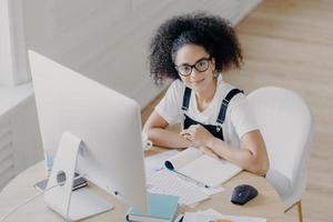 Top view of delighted curly female worker poses at desktop, dressed in casual wear, works on computer, makes financial report. Afro American student prepares for final exam in coworking space photo