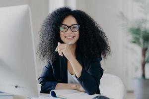 Positive international female student with crisp hair, wears transparent glasses, holds pen in hand, makes accountings, sits in front of big computer screen, dressed in black elegant outfit. photo