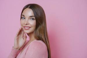 Photo of beautiful brown haired Caucasian woman keeps hand on cheek, smiles gently and looks directly at camera, has blue eyes, wears minimal makeup, stands in profile against pink background