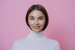 Isolated shot of charming glad young woman with pleasant smile, wears white turtleneck, looks directly at camera, isolated on pink background. Beautiful female student returns home from lectures photo