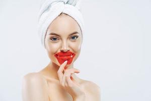 Pleasant looking healthy woman keeps patches on lips, wears towel on head, has clean pure skin after hygienic procedure, stands shirtless against white background, empty space for your advertisement photo