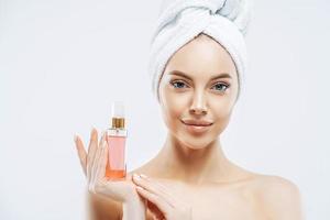 Portrait of charming young healthy woman holds bottle of expensive parfum, enjoys pleasant aroma, has healthy skin, wears bath towel on head, stands topless indoor. Women and cosmetics concept photo
