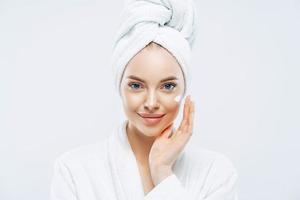 Young confident Caucasian woman applies face cream, enjoys new anti wrinkle cosmetic product, prevents sign of skin aging, wears minimal makeup, dressed in bath robe, isolated on white wall.