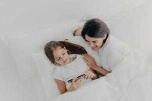 Shot of happy mother spends free time with little lovely daughter who is focused in smartphone, listens song in earphones, pose in bed with white soft bedclothes, have lazy morning during weekend photo