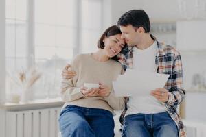Caring husband embraces wife with love, have glad expressions, holds paper documents, discuss banking procedures, study bills, drink tea or coffee, pose indoor, enjoy calm domestic atmosphere photo