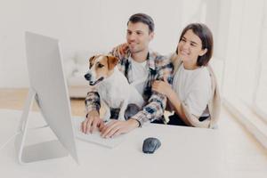Indoor shot of cheerful husband and wife look with joyful expressions, laugh as watch funny movie, rest together at free time, curious dog looks attentively at monitor of computer, man keyboards photo