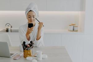 Young refreshed lady applies face powder with cosmetic brush looks at herself in mirror poses at kitchen at home sits at desk with beauty products wears wrapped towel and white dressing gown photo