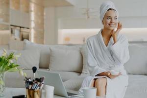 People beauty and skin care concept. Satisfied woman applies face cream enjoys facial treatments looks aside smiles gently uses cosmetic product poses at sofa in front of opened laptop computer photo
