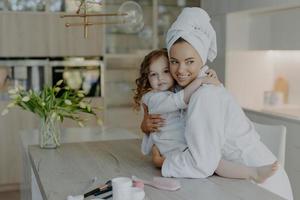 Photo of young mother and daughter embrace and express love to each other pose in modern apartment wear soft white bath robes going to have cosmetic procedures have healthy skin. Time for beauty