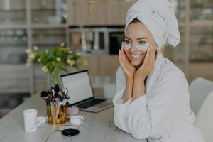 Pretty smiling young female model applies collagen patches under eyes to reduce wrinkles wears bathrobe and towel on head enjoys skin care procedures sits at desk with cosmetic tools cozy interior photo