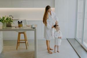 Caring mother combs hair of her small daughter wear white bathrobes pose against kitchen interior stand bare feet at floor. Beautiful little curly girl after taking shower. Mom making hairstyle to kid photo