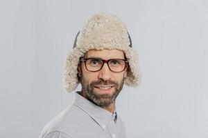 Close up shot of attractive male with stubble, wears glasses and fur hat, prepares for winter, has gentle smile models against white background. Masculine guy has blue eyes thick bristle stands indoor photo