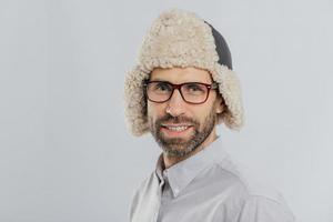 Photo of attractive young European male with pleasant appearance, wears warm fut hat, transparent glasses, models over white studio wall, rejoices shopping during weekend. People, style concept