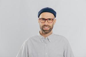 Horizontal view of pleased smiling man wears glasses, hat and shirt, being satisfied with good news, models against white background. Unshaven hipster rejoices new purchase. Fashion concept. photo