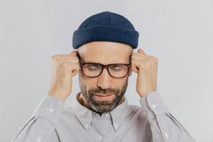 Close up shot of stressful unshaven man keeps hand near temples, has terrible headache, suffers from pain, wears headgear, spectacles, isolated over white background. Exhausted dissatisfied male photo