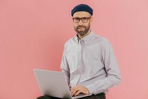 Male graphic designer thinks about creative ideas, reads news on internet website, uses moderm laptop computer, sits against pink background. Businessman reads publication online, makes research photo