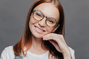 Close up portrait of pretty female model with European appearance, smiles gently, glad to recieve good comments from boss, wears spectacles, keeps hand near face, isolated over grey wall, tilts head photo