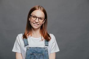 Isolated shot of positive pretty woman tilts head, has charming smile, wears glasses, white t shirt and overalls, poses over grey background with free space for your advertising content or promotion photo
