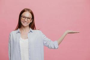 una mujer sonriente joven y positiva levanta la mano, finge sostener algo, gesticula sobre el espacio de la copia, vestida con una camisa de gran tamaño, usa anteojos, aislada sobre la pared rosa del estudio. anuncio publicitario foto