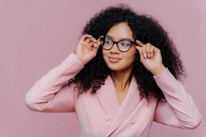 la foto de la atenta jefa mantiene las manos en el marco de las gafas, mira pensativamente hacia otro lado, usa un traje formal rosado, tiene un peinado afro rizado, posa contra un fondo morado. gente, negocios, etnia