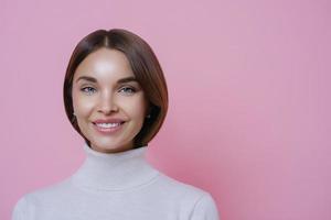 Close up portrait of lovely satisfied joyful woman with dark hair, smiles pleasantly, shows white teeth, wears poloneck, isolated on pink background, empty space for your advertising content. photo