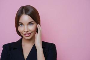 Photo of lovely European female entrepreneur with healthy skin, touches face gently, returns from spa salon, wears formal black outfit, looks confidently at camera, isolated on pink background