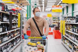 Horizontal shot of man walks in supermarket with shopping trolley with goods, wears medical mask and rubber gloves, food running out because of coronavirus. Virus outbreak. Panic buying concept. photo