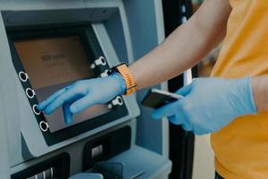 Cropped shot of man wears protective rubber gloves, uses ATM machie to withdraw money, prevents from corona virus or covid-19, holds mobile phone, presses button. Virus pandemic. Health care concept photo