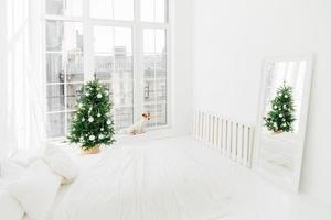 New Year interior. Spacious white bedroom with bed, decorated Christmas tree, mirror and jack russell terrier dog on windowsill. Pet waits for Santa Claus photo