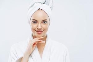 Cropped shot of young good looking European woman looks with calm face, enjoys bath procedures, wears soft white robe, wrapped towel on head, isolated on white background. Morning time, hygiene photo