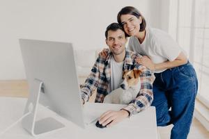 Indoor shot of happy family couple pose near computer monitor, work together remotely, have own business, earn money online, look gladfully at camera, play with favourite pet. People, technology photo