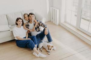 Lovely European family of father, mother and their daughter sit on floor near sofa in spacious white room, pedigree dog lies near, have moving day, change place of living, express happiness. photo