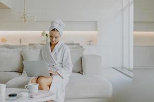 Pleased young female model concentrated at laptop screen wears bathrobe and towel on head poses in spacious living room at sofa enjoys spending weekend at home feels comfortable and relaxed. photo