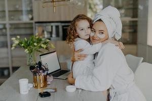 Attractive woman wears hydrogel patches under eyes dressed in bathrobe and wrapped towel embraces her small daughter with love pose together at home near cosmetic tools. Makeup routine concept photo