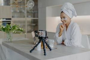 Beautiful successful female blogger records tutorial video talks with followers looks at camera of smartphone on tripod applies beauty patches under eyes dicuss how to care about skin and complexion photo