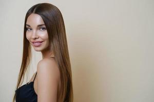 Portrait of pleased delighted woman with long dark hair, smiles gently, looks with attentive gaze at camera, poses against beige background, blank copy space aside. Party preparation, femininity photo