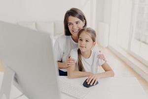 Happy affectionate mom embraces her small daughter, work together on modern computer, preapare presentation for lesson, look gladfully into monitor, spend leisure time at home, surf internet page. photo