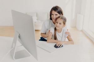 Modern technology and family concept. Caring lovely mother teaches little daughter how to use internet, explains how to download application, pose in spacious room with white walls, spend time at home photo