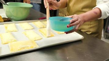 primo piano delle mani di uno chef con grembiule cappotti creme caramello dai tuorli d'uovo in pasta sfoglia con un pennello prima di cuocere una torta e cibi da forno in una cucina di corso di cucina culinaria. video