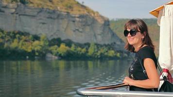 Brunette woman in black t-shirt enjoying seascape, standing on ferry deck at sunny day. Pretty girl in sunglasses looking away, smiling, with cliff on background. Concept of travel video