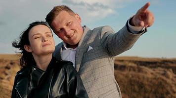 Portrait view of man in suit showing view to pretty woman, carissing her while standing on hill at sunset. Happy, young couple kissing, cuddling with blue sky on background. Concept of romance video