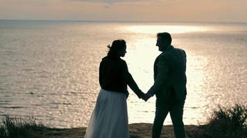 Back view of wedding couple wearing casual clothes walking on seashore toward setting sun. Bride and groom walking, holding hands, hugging with panoramic view of ocean at sunset. Concept of wedding video