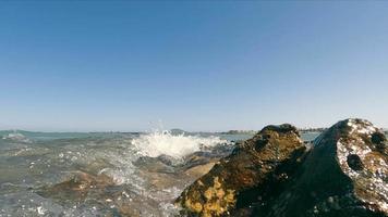 vista de cerca de las olas del mar rompiendo en las rocas costeras en un día soleado sin nubes. cámara lenta de olas burbujeantes, rodando en la costa rocosa con cielo azul claro en el fondo, espacio de copia. concepto de paisaje marino video