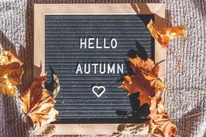 Autumnal Background. Black letter board with text phrase Hello Autumn and dried leaves lying on white knitted sweater. Top view, flat lay. Thanksgiving banner. Hygge mood cold weather concept photo